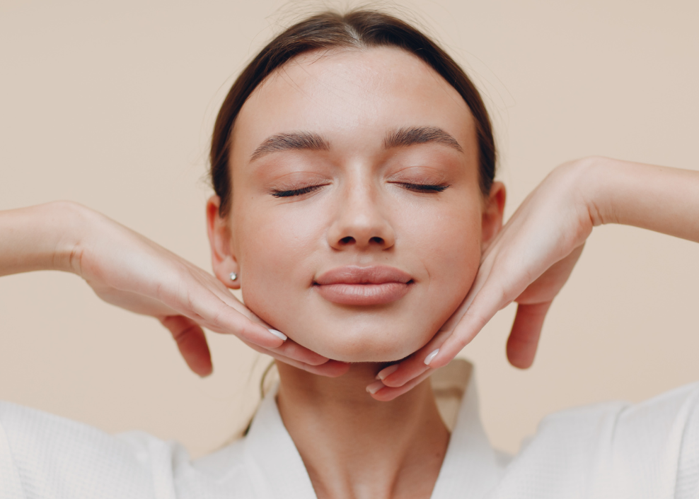 Woman in a white robe with her eyes closed, hands on her face against a beige background